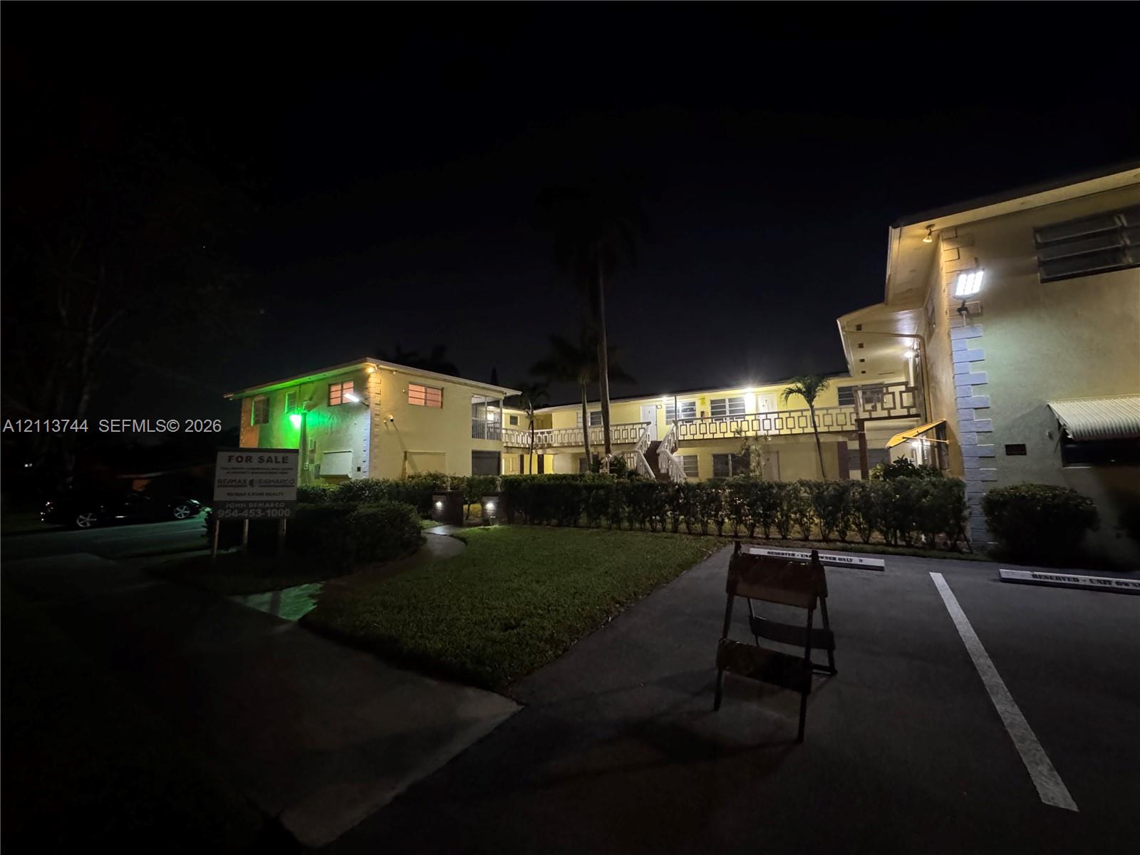 Multi-story exterior featuring stucco siding, metal railing balconies, paved parking area, and mature palm trees