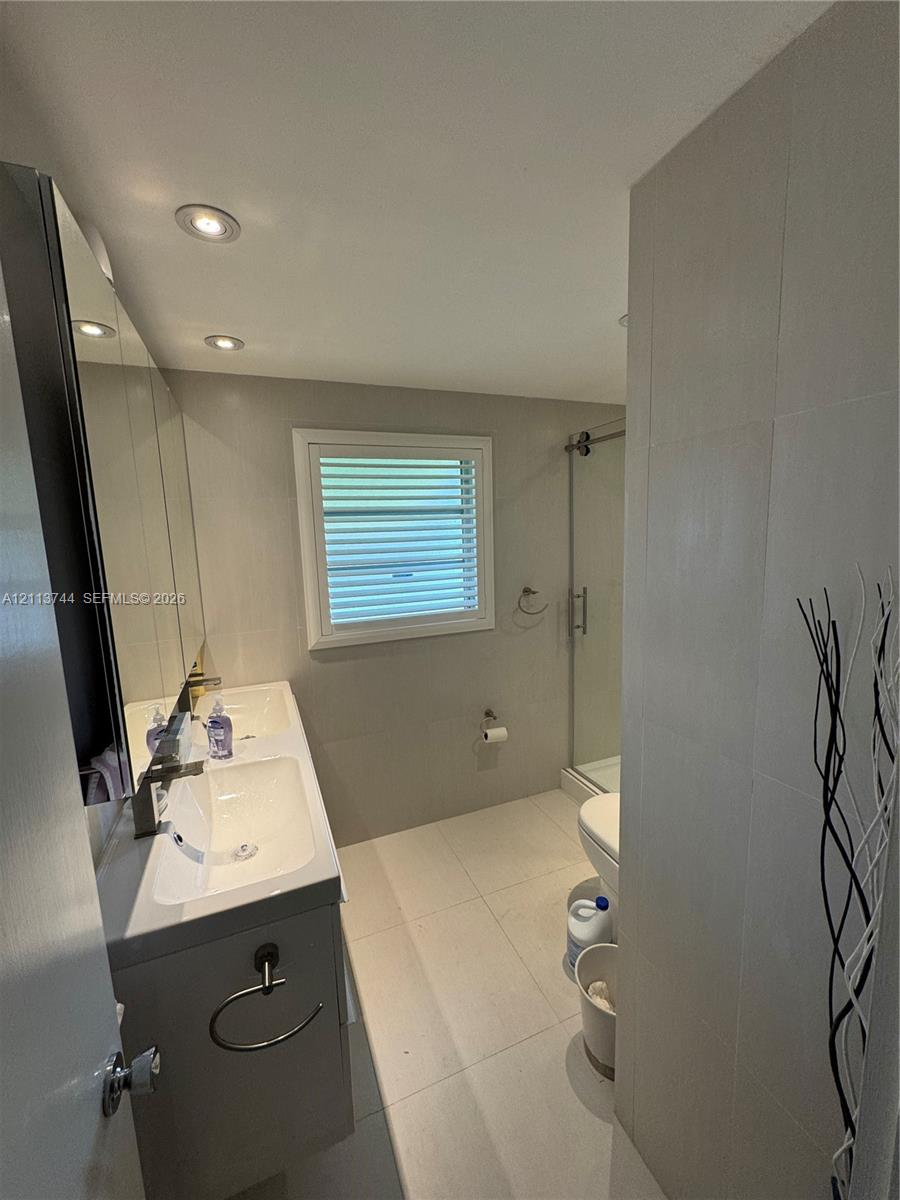 Dual-sink vanity with a rectangular countertop, large-format tile flooring, wall-to-wall mirror, recessed lighting, and a glass-enclosed shower