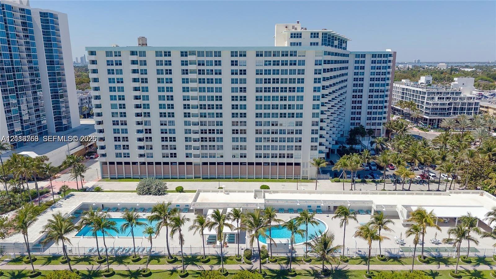 High-rise residential exterior featuring a multi-story facade, dual ground-level swimming pools, paved pool deck, and mature palm tree landscaping