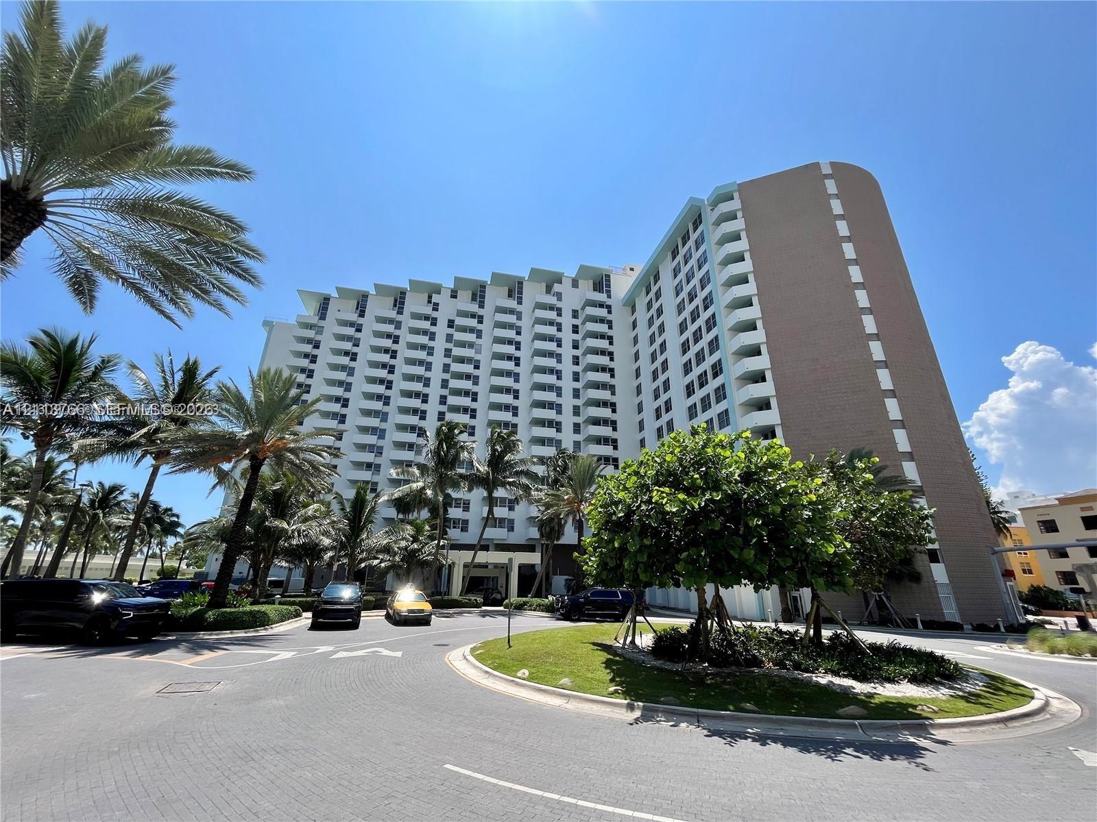 High-rise residential exterior featuring multiple tiers of private balconies, architectural concrete facade, circular paved motor court, and mature tropical landscaping