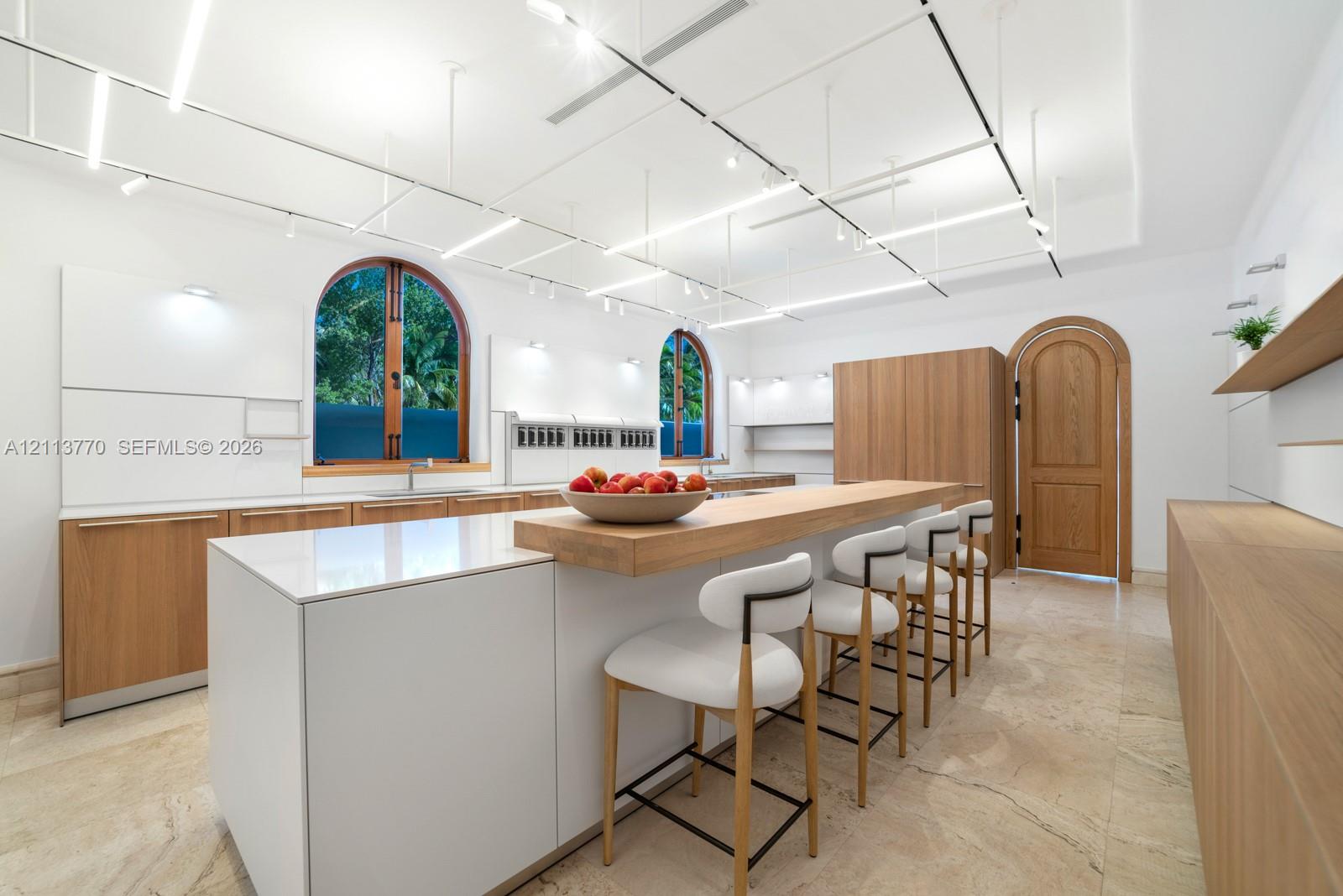 Kitchen featuring flat-panel cabinetry with wood-finish and matte-white surfaces, central island with an integrated wood-grain breakfast bar, arched windows, stone-tile flooring, and grid-pattern overhead track lighting