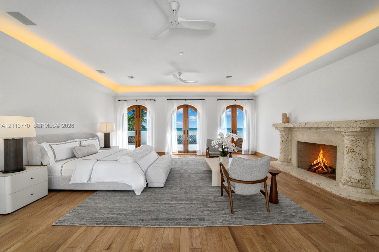 Spacious room featuring wood-finish flooring, recessed tray ceiling lighting, dual white ceiling fans, and an ornate, textured stone fireplace surround with arched French doors