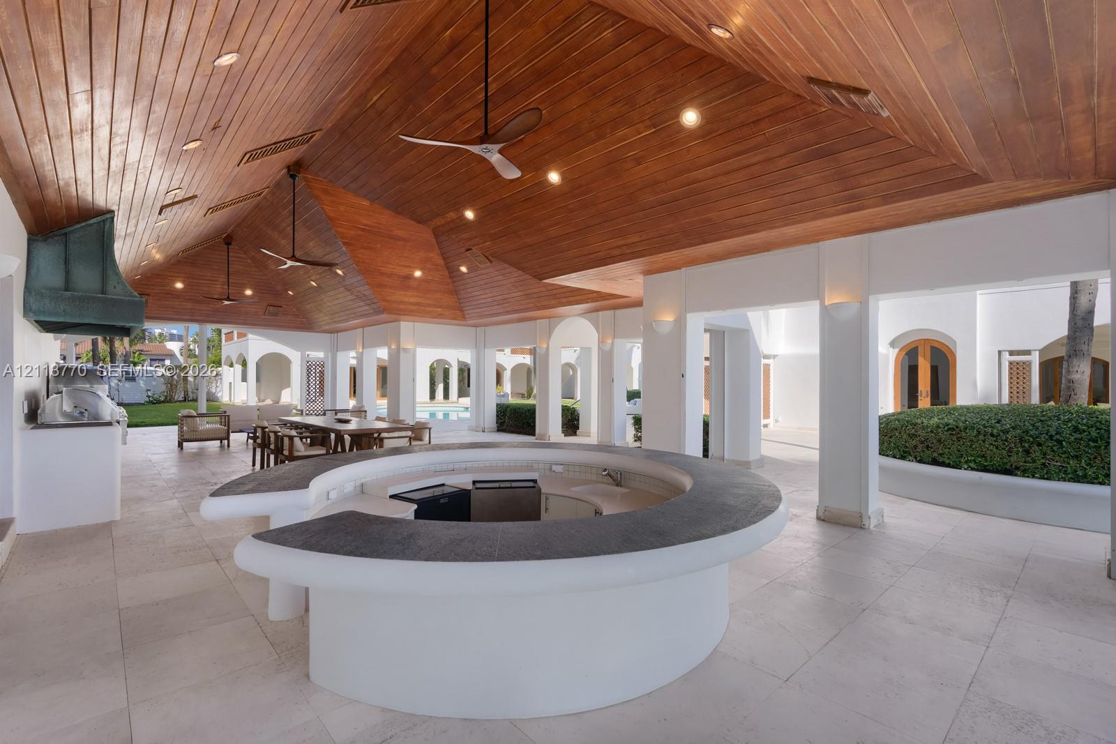 Covered outdoor patio featuring a vaulted tongue-and-groove wood ceiling, a circular concrete bar with a dark stone-finish countertop, stone flooring, and built-in recessed lighting