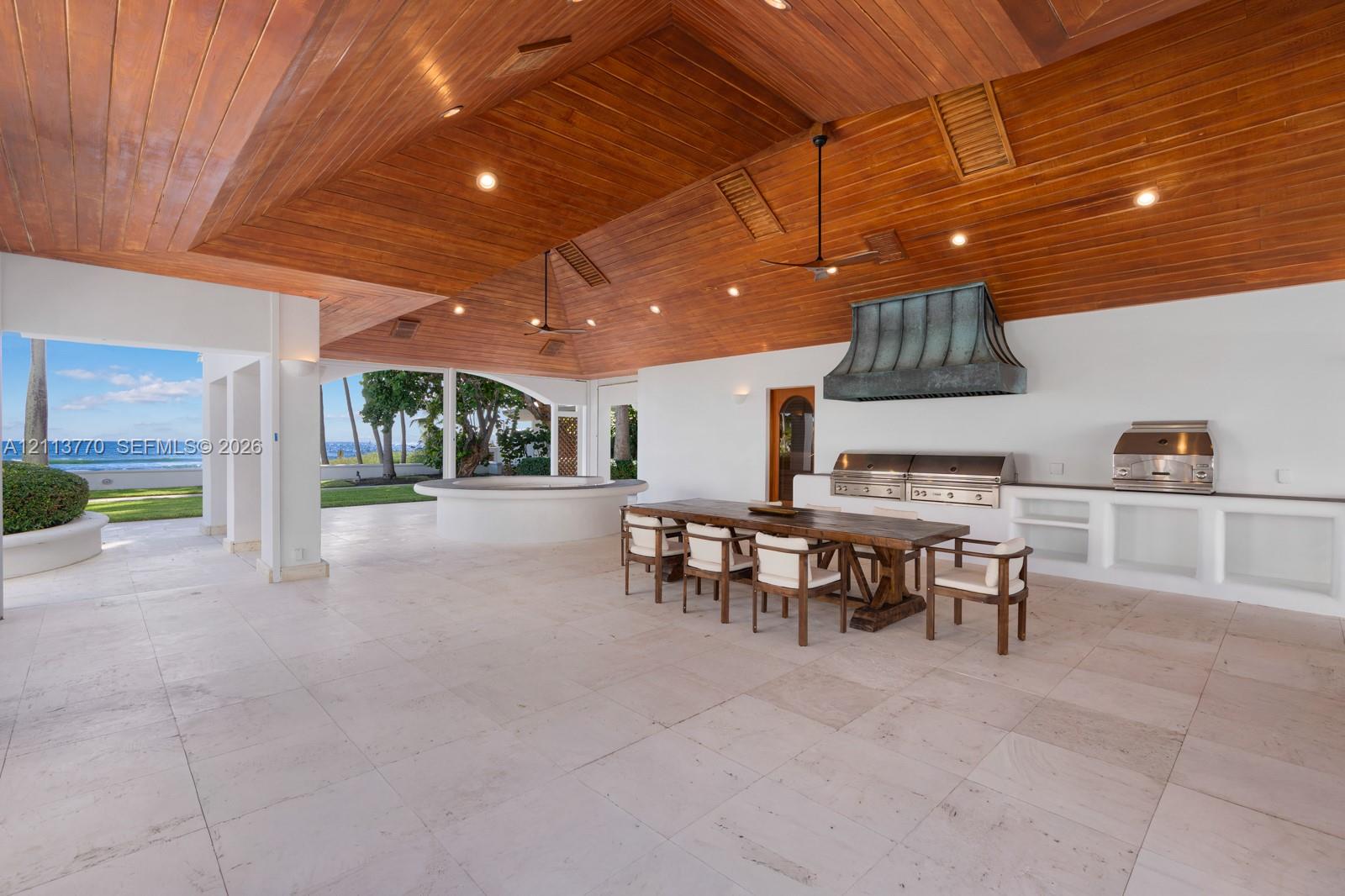 Covered outdoor kitchen featuring vaulted wood-paneled ceilings, stone flooring, built-in stainless steel appliances, and a custom metal range hood