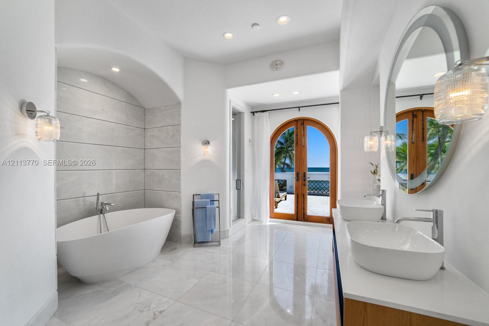 Freestanding soaking tub set within a tiled arched alcove, dual oval vessel sinks with polished chrome faucets, quartz countertop vanity, and arched double doors featuring exterior ocean scenery