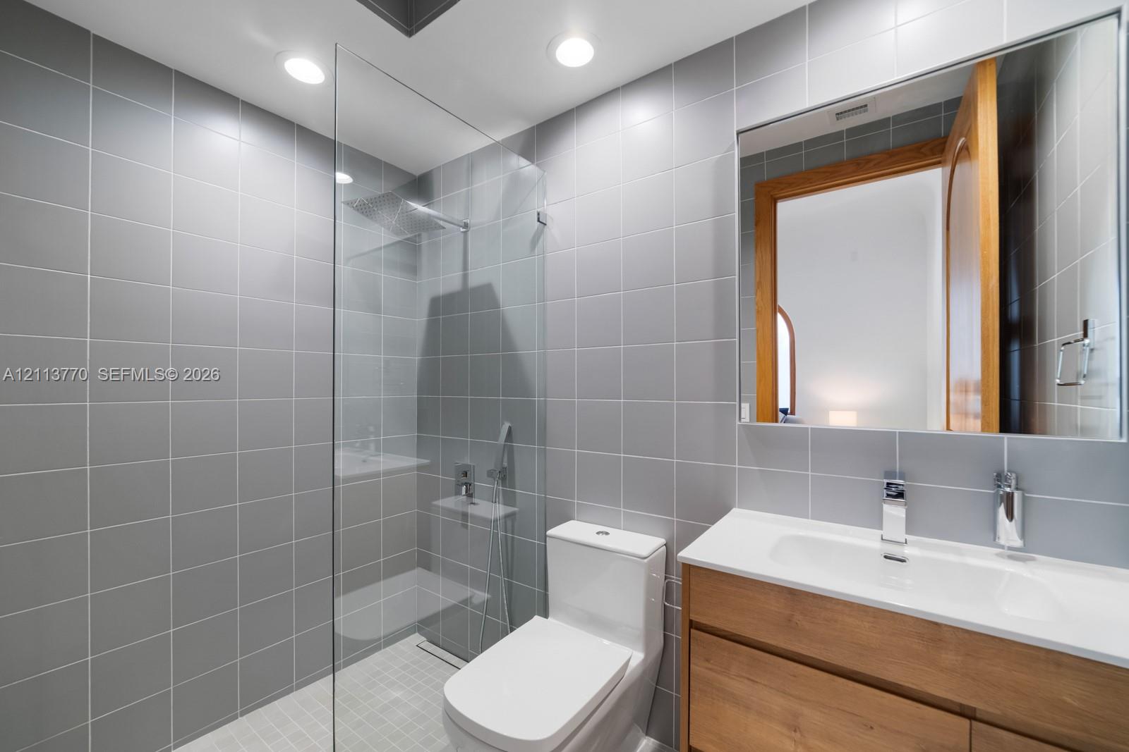 Walk-in shower with a glass panel and rainfall showerhead, vanity with a white countertop and integrated sink, and floor-to-ceiling gray wall tiling