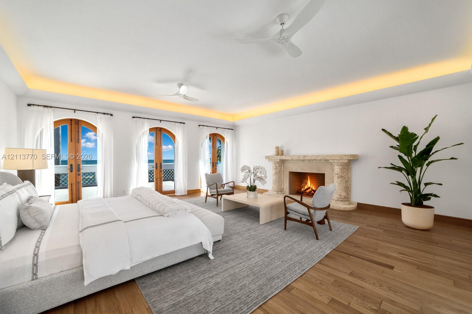 Spacious room featuring wood-finish flooring, recessed tray ceiling lighting, three sets of arched wooden-framed glass doors, and a limestone-surround fireplace with decorative columns