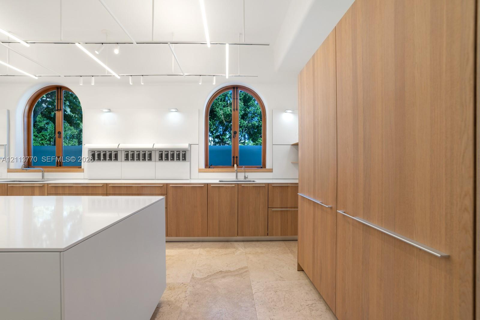 Kitchen featuring light-finish wood cabinetry, a solid-surface center island, arched windows with wood frames, stone tile flooring, and integrated linear overhead lighting