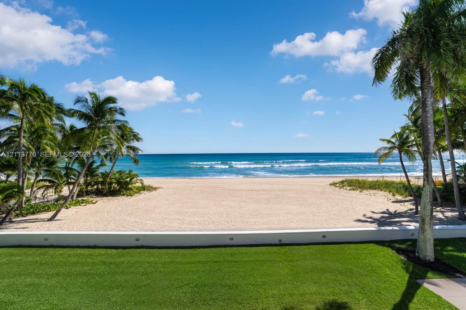 Direct beach access, manicured lawn, low-profile concrete retaining wall with integrated lighting, and mature palm trees