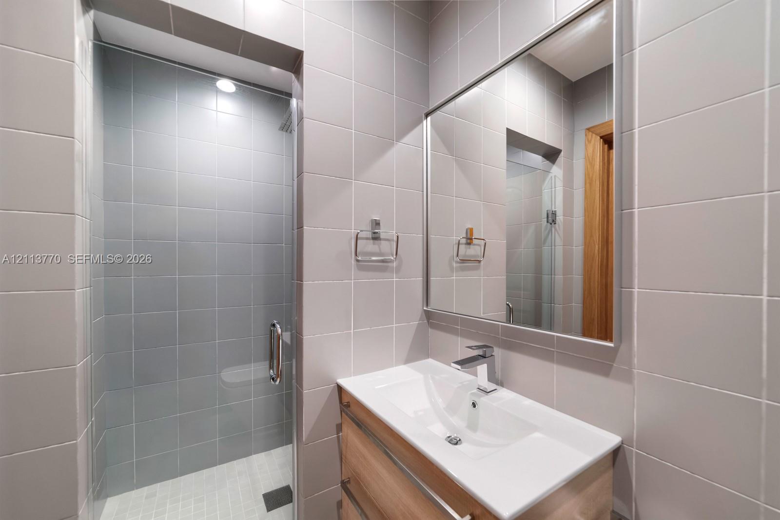 Bathroom featuring a light wood-finish floating vanity with a white integrated sink, chrome single-lever faucet, wall-to-wall square ceramic tiling, and a glass-enclosed shower