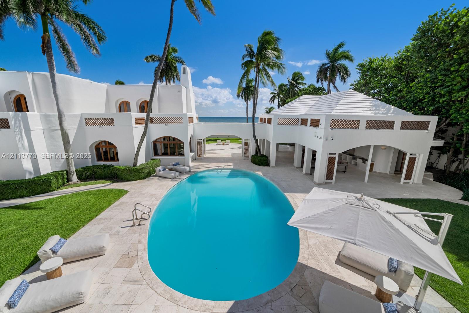 Stucco exterior featuring arched architectural detailing, a central swimming pool with stone-tiled decking, a covered outdoor loggia, and integrated privacy hedging