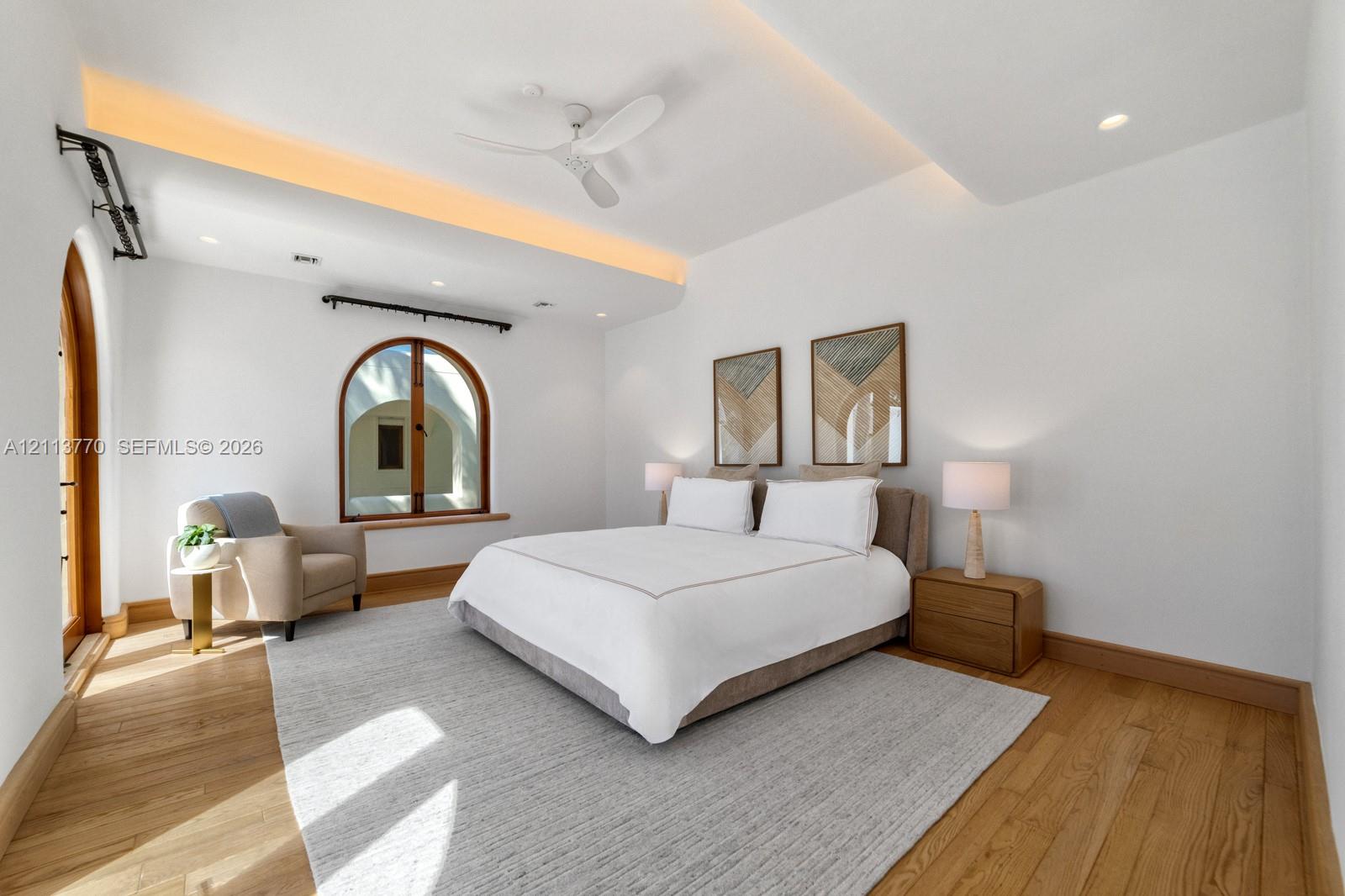 Room featuring wood-finish flooring, tray ceiling with recessed accent lighting, ceiling fan, and arched window and door frames with wood casing