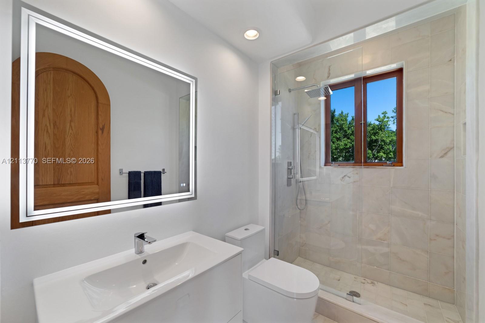 Full bathroom featuring a glass-enclosed shower with tiled surround, light-toned stone-finish flooring, a wall-mounted LED mirror, and a white vanity with an integrated basin