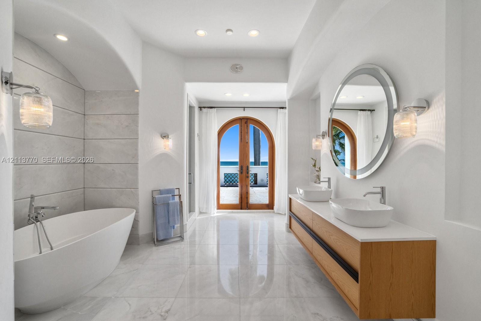 Freestanding soaking tub set within a tiled arched alcove, dual-sink floating vanity with white surface, round wall-mounted mirror, recessed lighting, and arched wood-frame glass doors