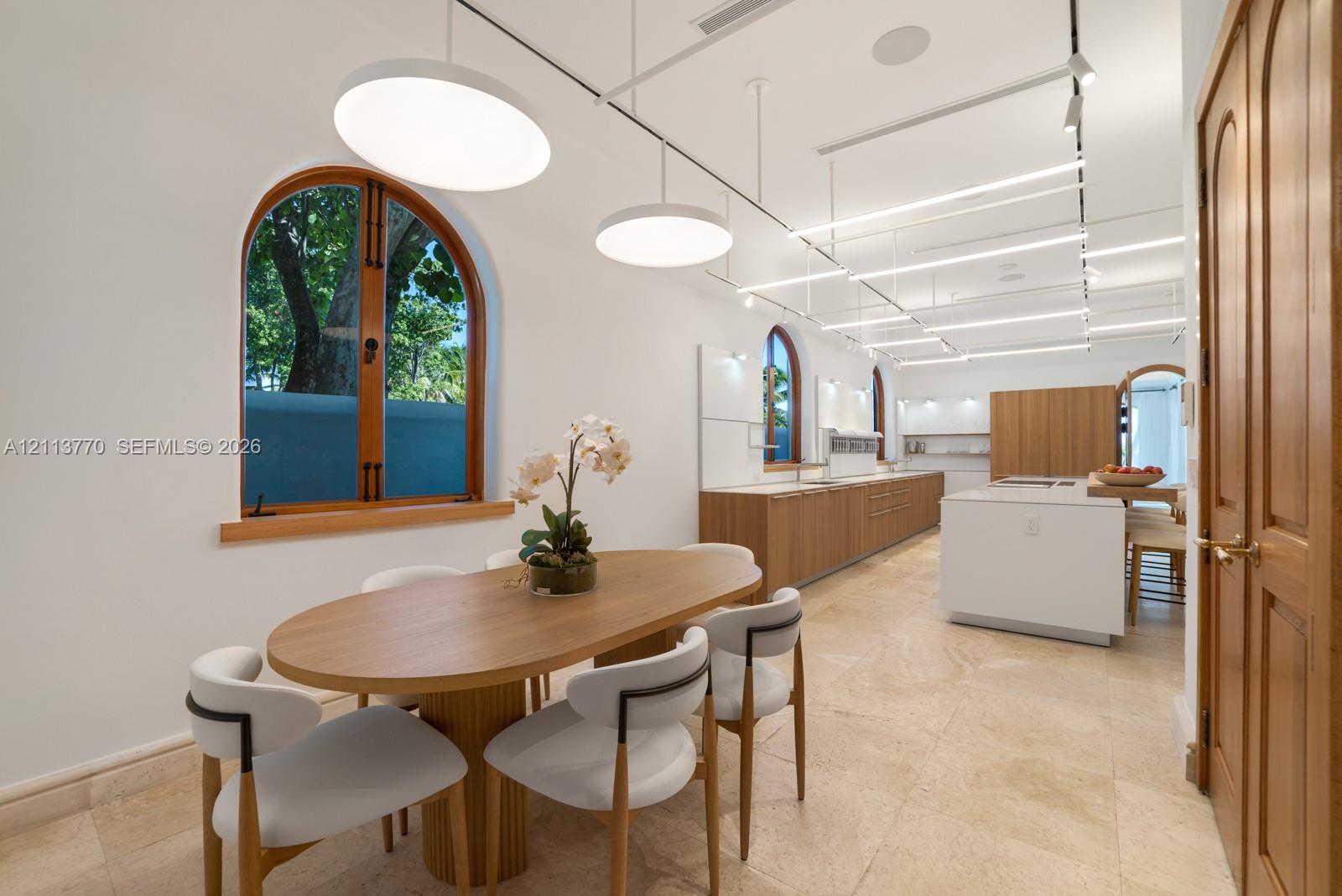 Open-concept space featuring arched wood-framed windows, stone flooring, integrated linear track lighting, and a kitchen island with built-in cabinetry