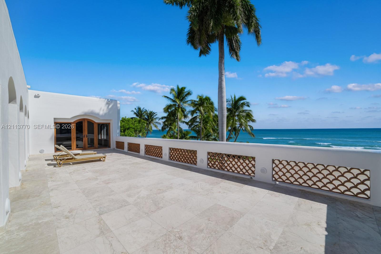 Expansive stone terrace featuring decorative metal balustrades, arched wood-framed glass double doors, and ocean frontage