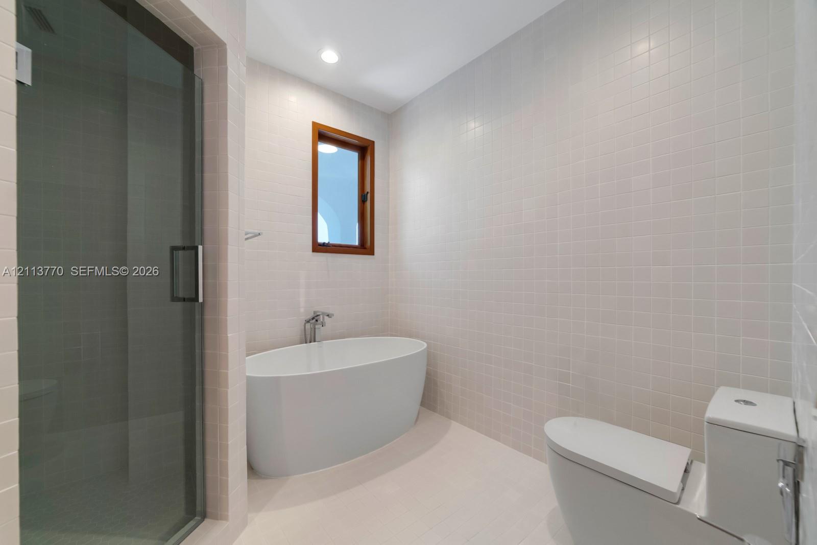 Freestanding soaking tub with wall-mounted faucet, glass-enclosed walk-in shower, floor-to-ceiling white tile surround, and wood-framed window