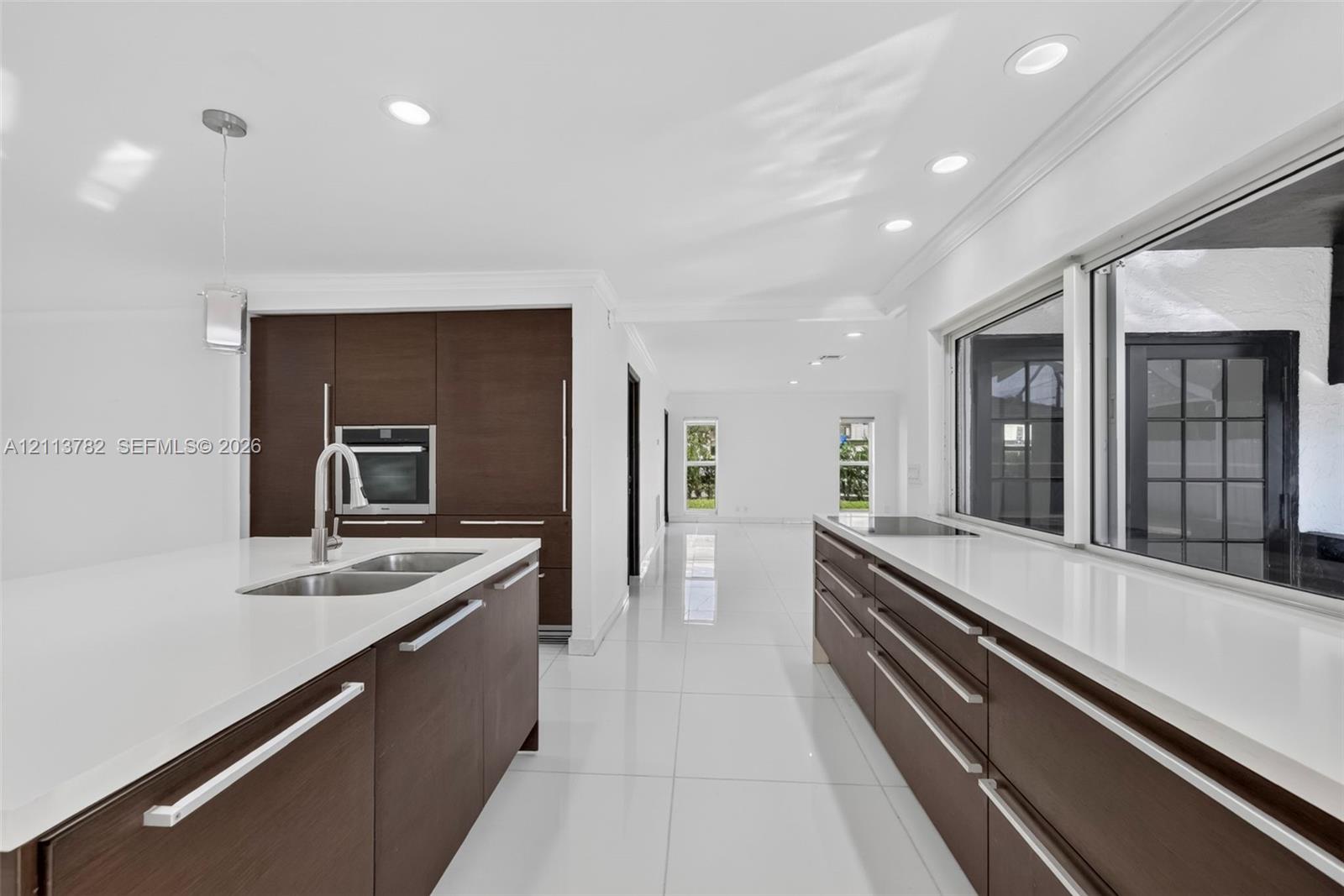 Kitchen featuring dark wood-finish cabinetry, solid-surface countertops, integrated wall oven, stainless steel dual-basin sink with pull-down faucet, and polished tile flooring