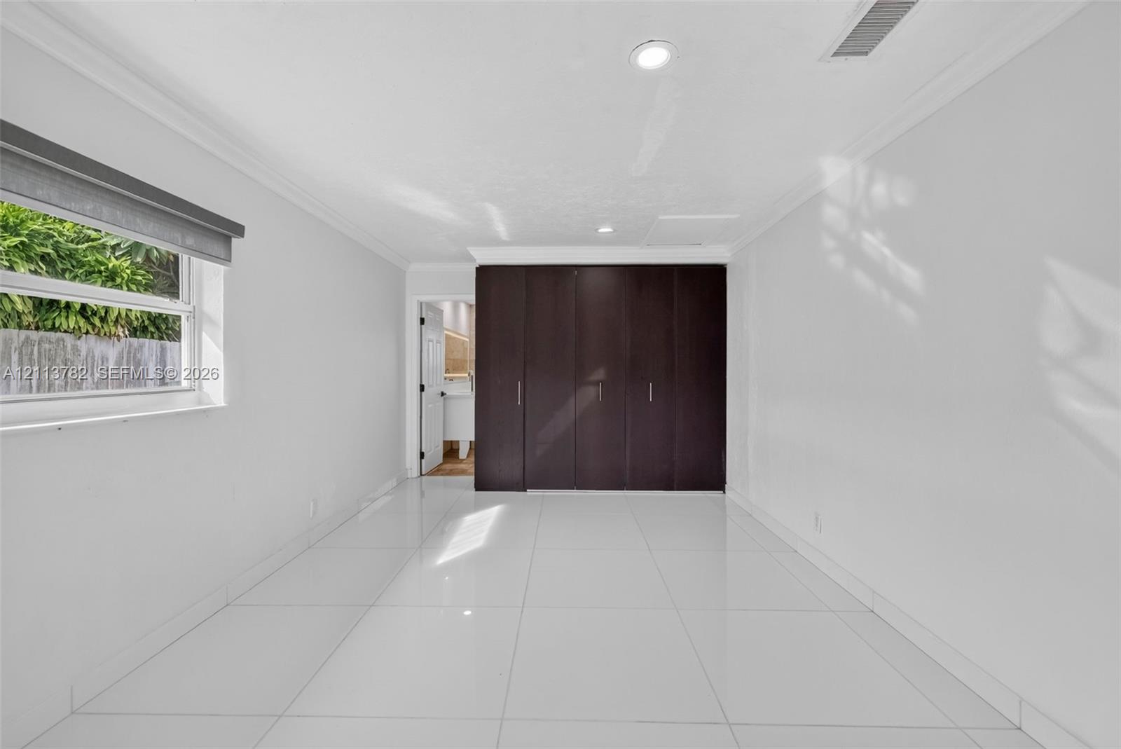 Neutral-toned interior featuring large-format white tile flooring, crown molding, recessed lighting, and floor-to-ceiling built-in cabinetry with dark wood-finish doors