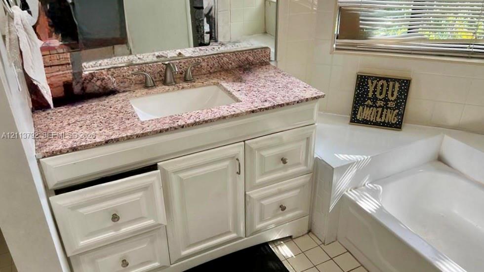 Bathroom vanity featuring white cabinetry, a stone-composite countertop, and a recessed sink basin paired with a soaking tub and square floor tile