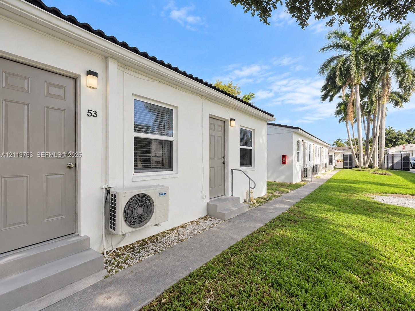 Stucco exterior featuring minimalist entryways, dark-toned panel doors, white-framed windows, wall-mounted exterior lighting, and an expansive lawn with mature palm trees Stucco exterior featuring minimalist entryways, dark-toned panel doors, white-framed windows, wall-mounted exterior lighting, and an expansive lawn with mature palm trees