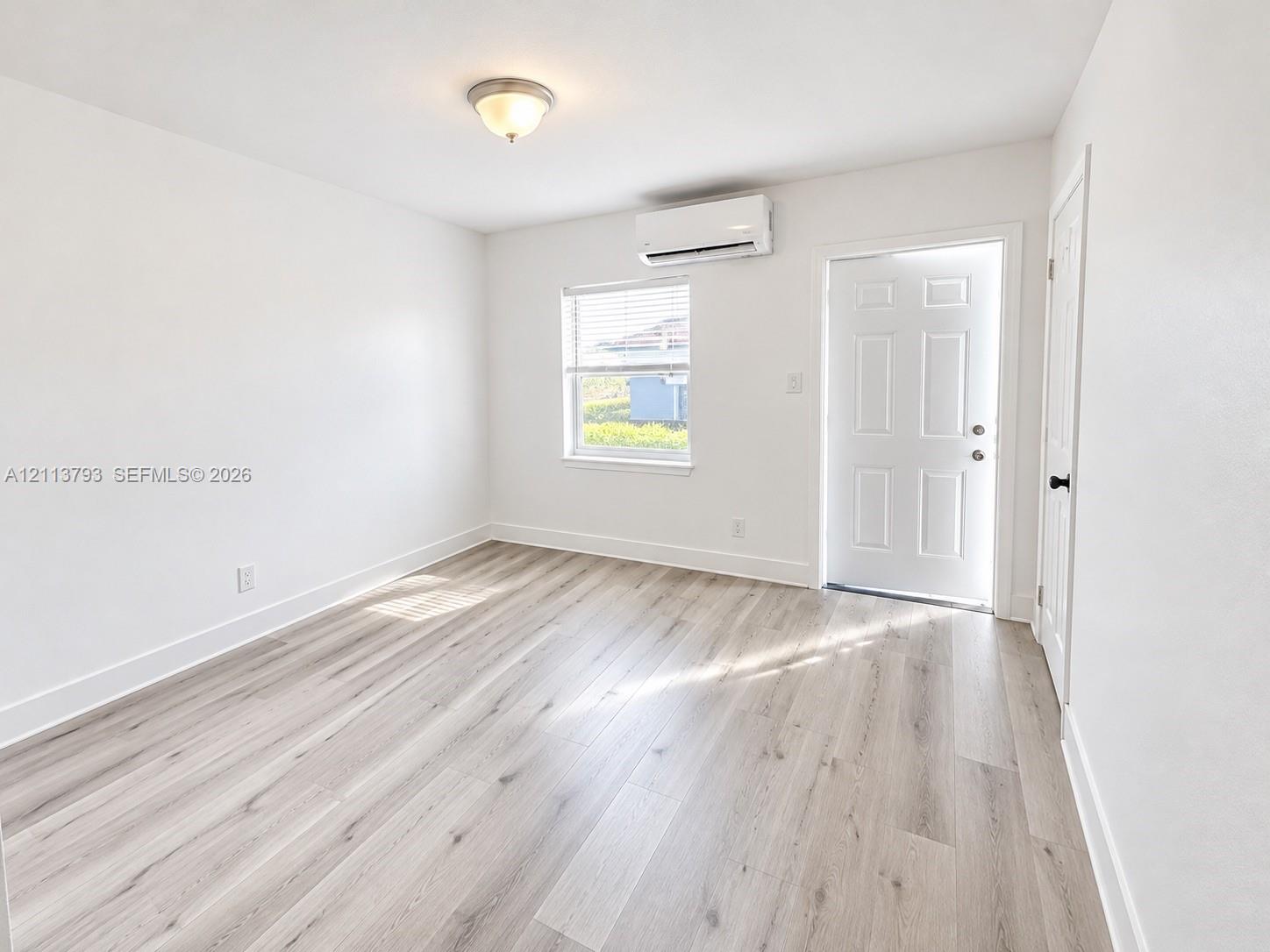 Interior space featuring wood-finish flooring, a wall-mounted ductless mini-split air conditioning unit, a six-panel exterior entry door, and a single-hung window with blinds Interior space featuring wood-finish flooring, a wall-mounted ductless mini-split air conditioning unit, a six-panel exterior entry door, and a single-hung window with blinds