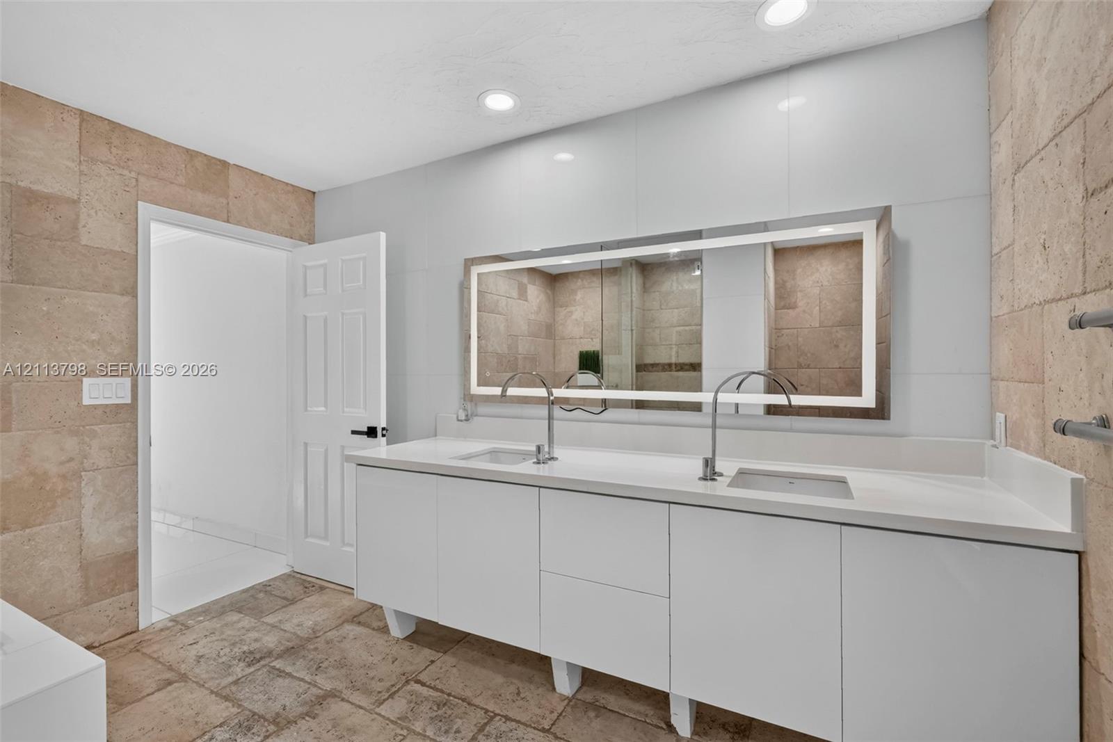 Double vanity with white cabinetry, solid surface countertop, rectangular undermount sinks, tall polished metal faucets, and a large backlit wall-mounted mirror surrounded by textured stone wall tile