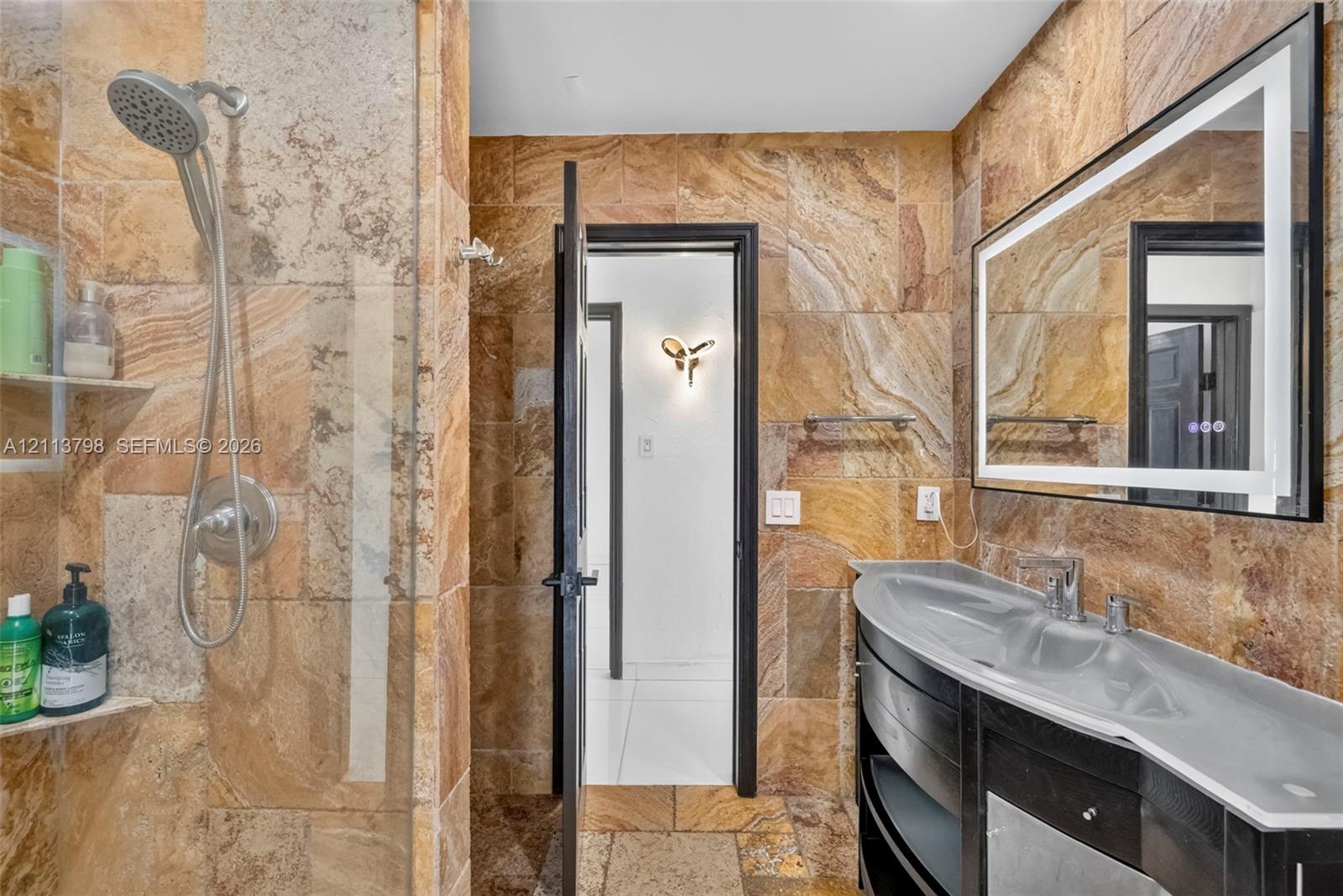 Bathroom featuring natural stone tile wall and floor surfaces, a curved vanity with a basin sink, an integrated-lighting rectangular mirror, and a wall-mounted shower head