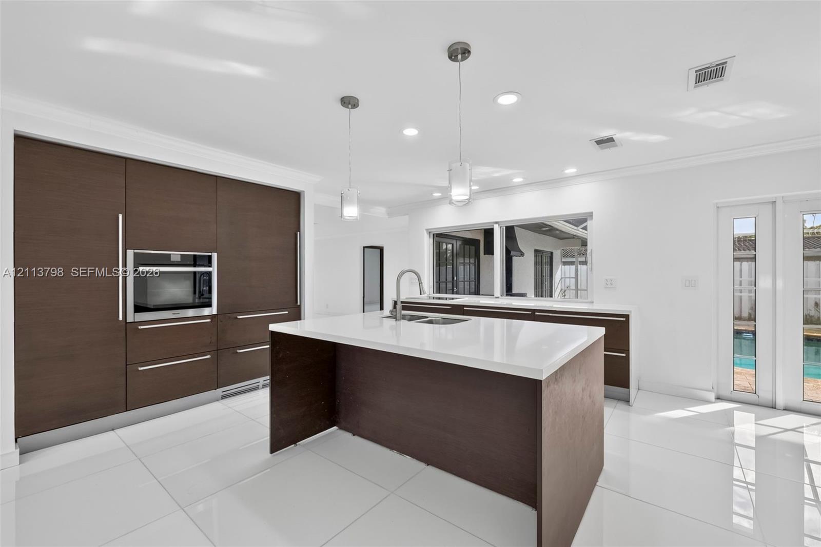 Kitchen featuring dark wood-finish cabinetry, solid-surface countertops, a center island with a built-in sink, stainless steel wall oven, and polished tile flooring Kitchen featuring dark wood-finish cabinetry, solid-surface countertops, a center island with a built-in sink, stainless steel wall oven, and polished tile flooring