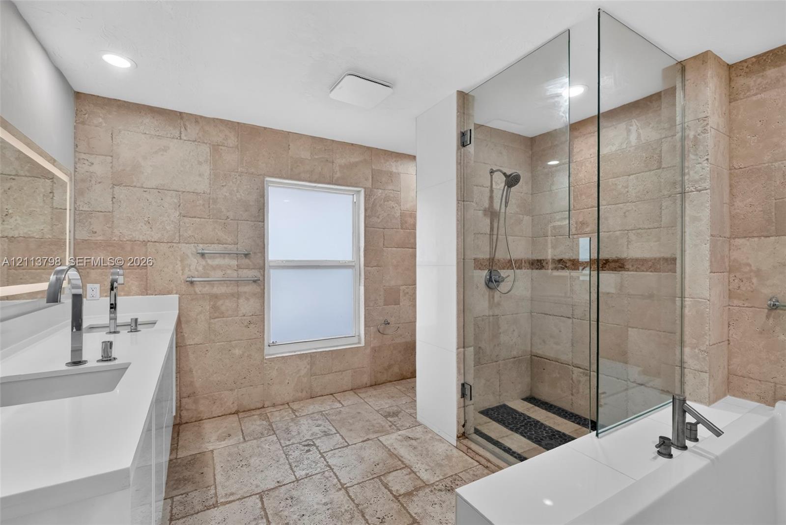 Bathroom featuring natural stone wall and floor tiling, double-basin vanity with solid-surface countertop, glass-enclosed walk-in shower with rainfall showerhead, and a deep soaking tub