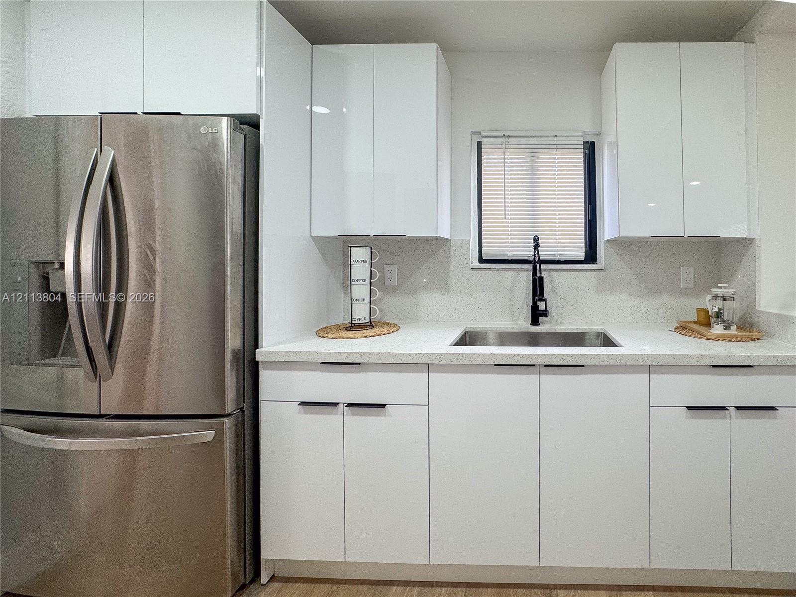 Kitchen featuring white high-gloss cabinetry, stainless steel French door refrigerator, light-toned solid surface countertops, undermount sink, and matte black high-arch faucet Kitchen featuring white high-gloss cabinetry, stainless steel French door refrigerator, light-toned solid surface countertops, undermount sink, and matte black high-arch faucet