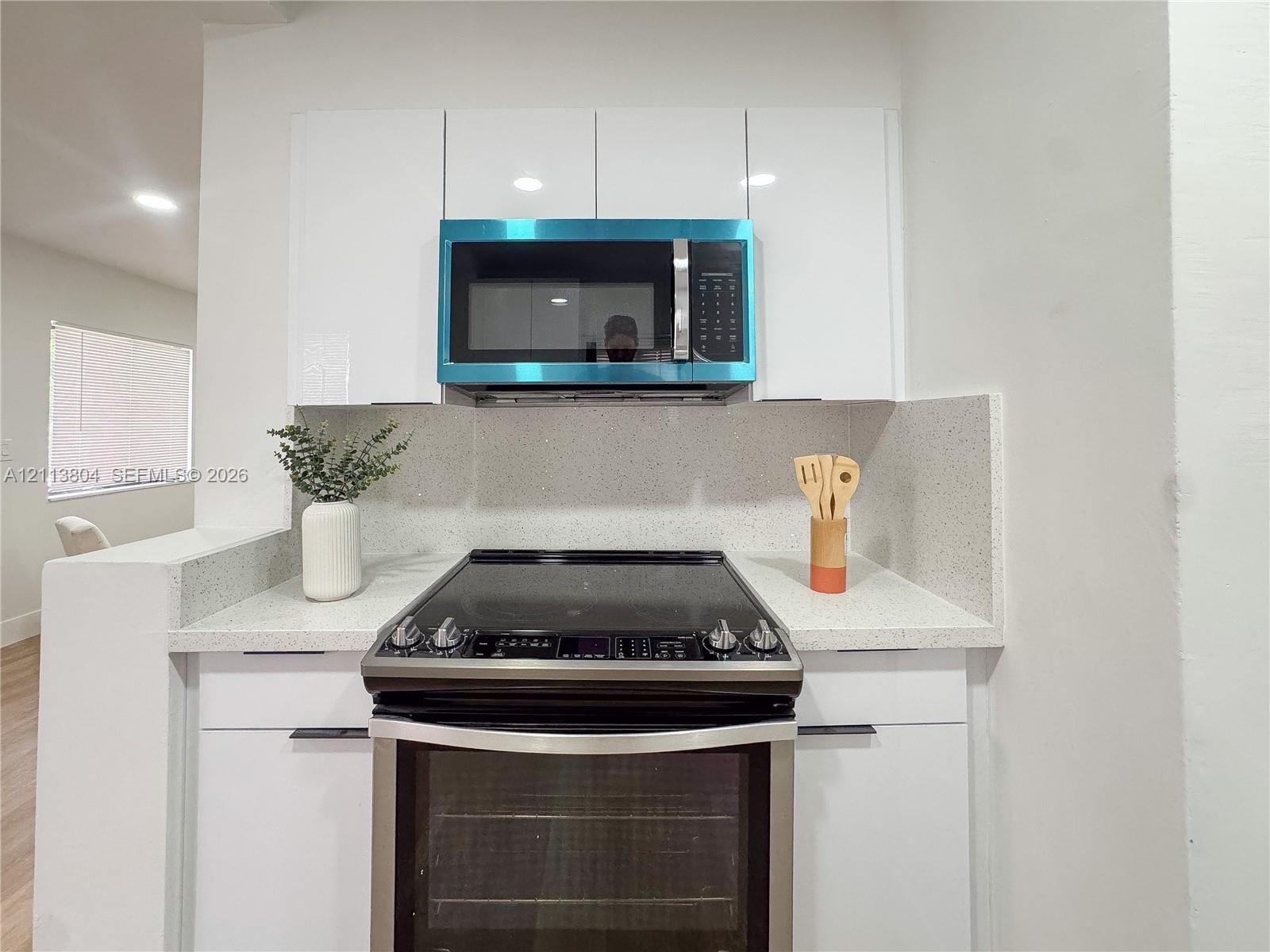 Gloss-finish white cabinetry featuring flat-panel doors, stone-composite countertops, high-back matching backsplash, stainless steel electric range, and over-the-range microwave Gloss-finish white cabinetry featuring flat-panel doors, stone-composite countertops, high-back matching backsplash, stainless steel electric range, and over-the-range microwave