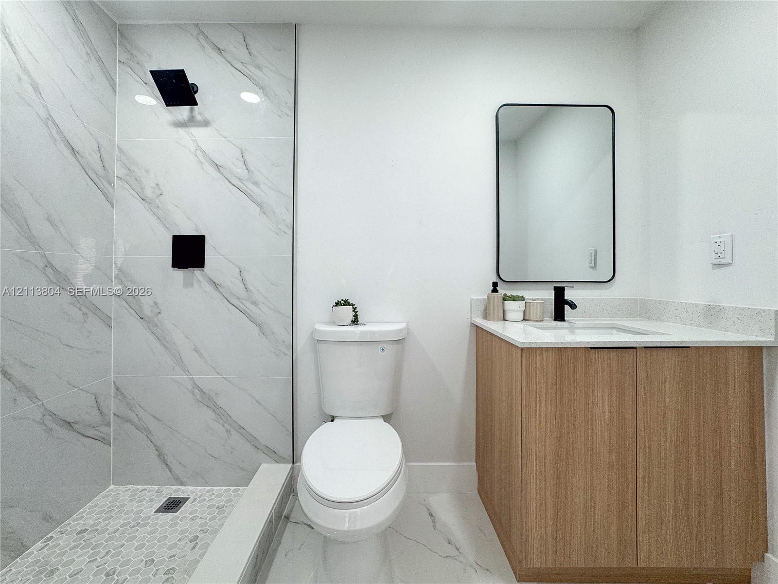 Full-height shower surround with veined stone-finish tile, pebble-tile shower floor, matte black shower fixture, light wood-finish vanity with stone-finish countertop, and wall-mounted rectangular mirror with a black frame