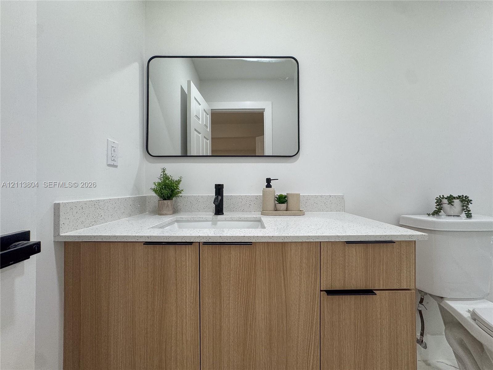 Wood-finish cabinetry featuring black hardware, quartz-composite countertop, undermount sink, matte black faucet, and rectangular black-framed vanity mirror