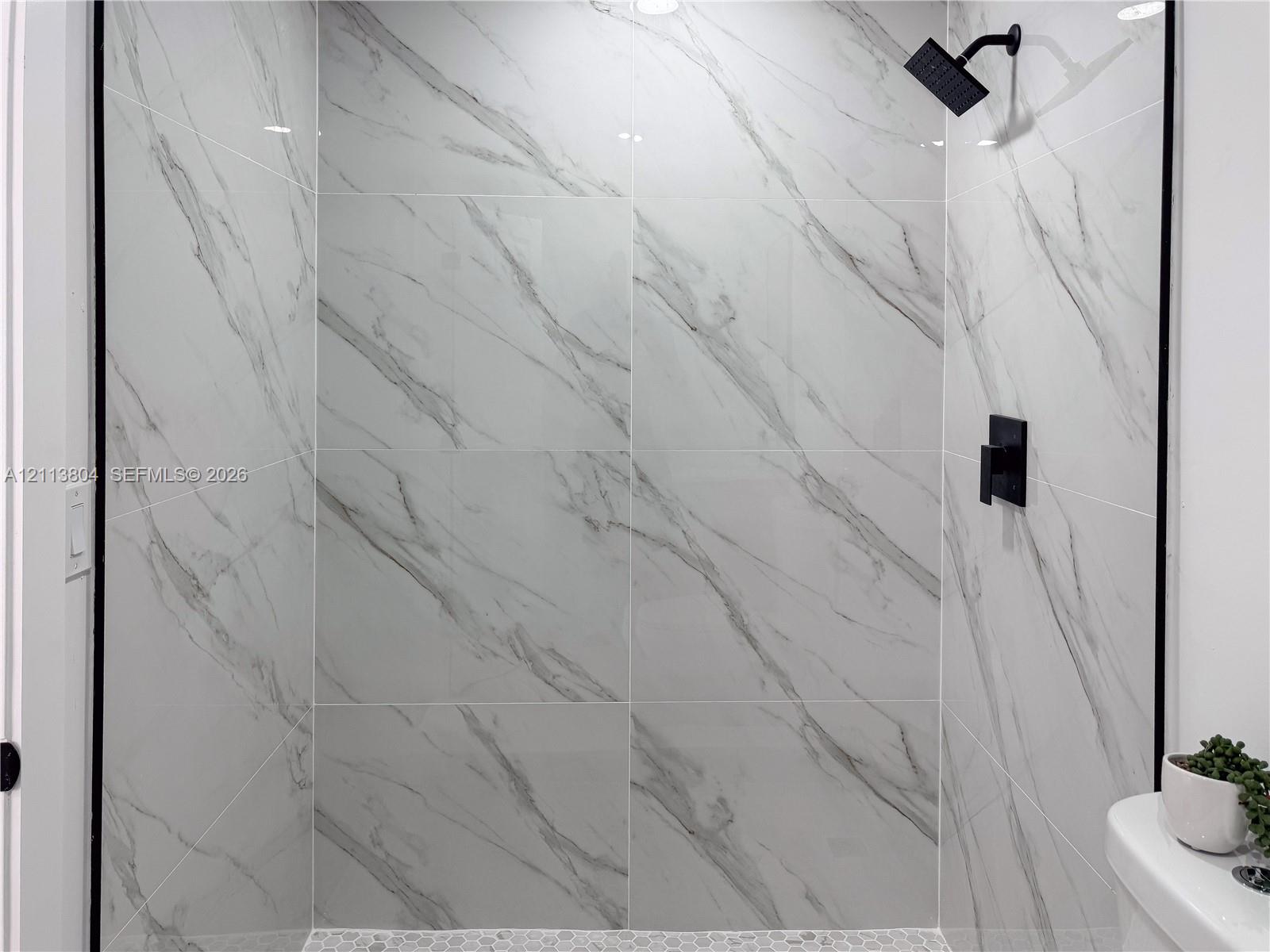 Shower featuring floor-to-ceiling large-format tile with decorative veining, matte black wall-mounted shower head, matte black single-handle mixing valve, and stone hexagon mosaic floor tile