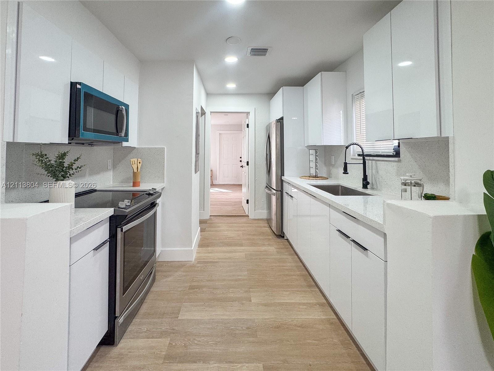 Kitchen featuring high-gloss white cabinetry, white stone-finish countertops, stainless steel appliances, wood-finish flooring, and a matte black gooseneck faucet Kitchen featuring high-gloss white cabinetry, white stone-finish countertops, stainless steel appliances, wood-finish flooring, and a matte black gooseneck faucet