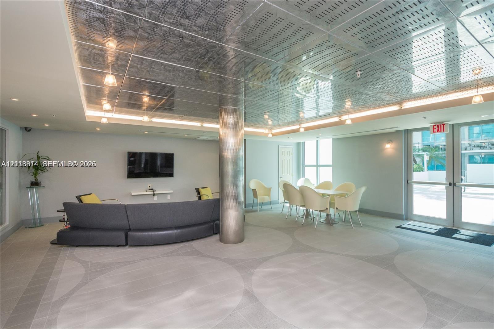 Metallic paneled ceiling with integrated cove lighting, central metallic support column, patterned tile flooring, and glass-paned double entry doors