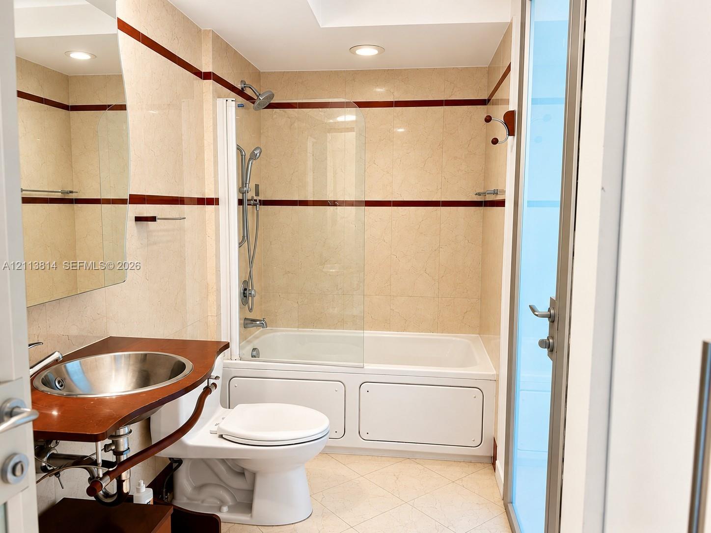 Bathroom featuring full-height neutral wall tiling with dark accent banding, a wood-surround vanity with a circular metallic vessel sink, a white drop-in soaking tub, and a glass shower panel