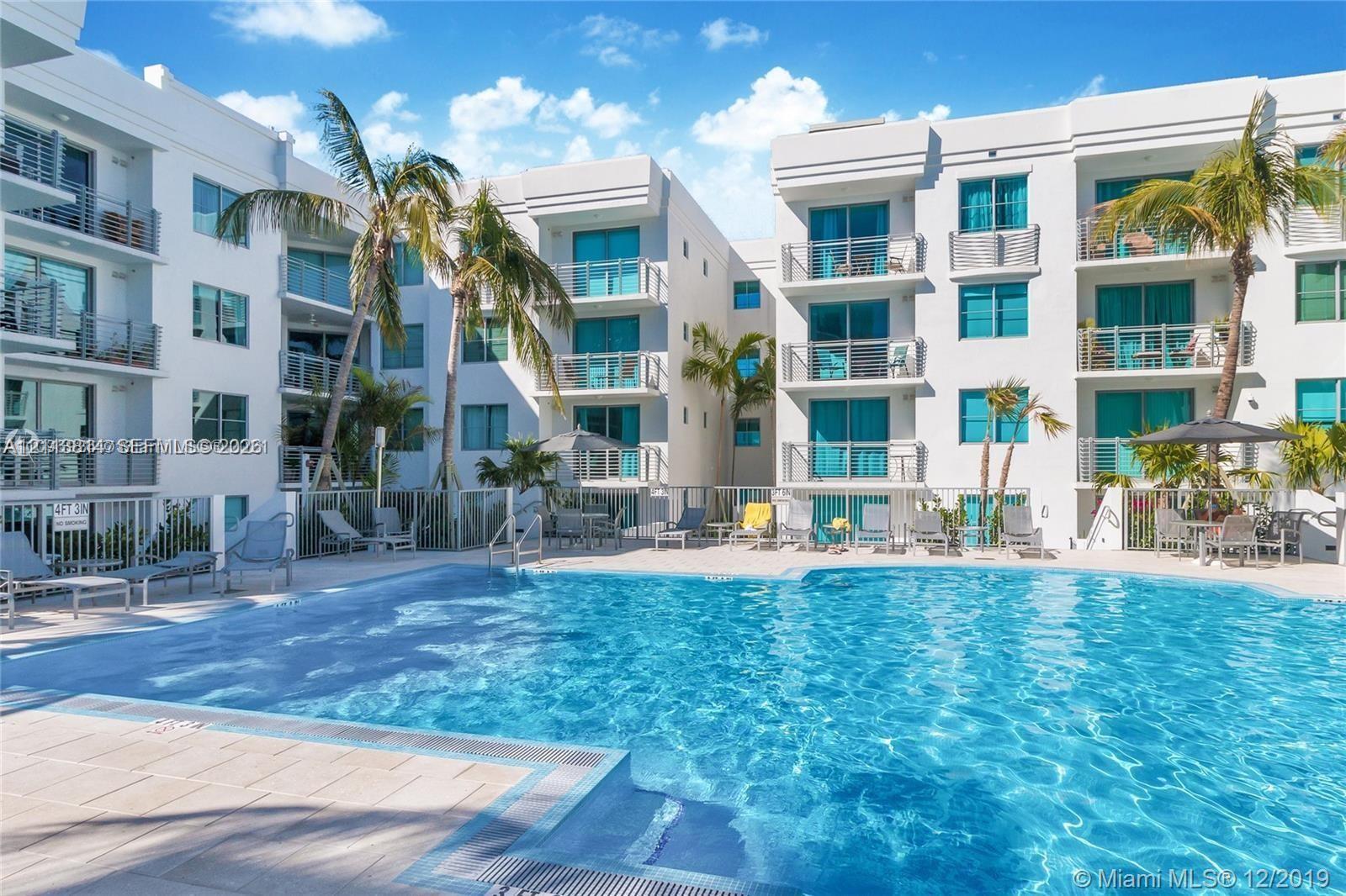 Swimming pool with light-colored tile decking, surrounded by multi-story white stucco building facades featuring private balconies with metal railings and palm landscaping