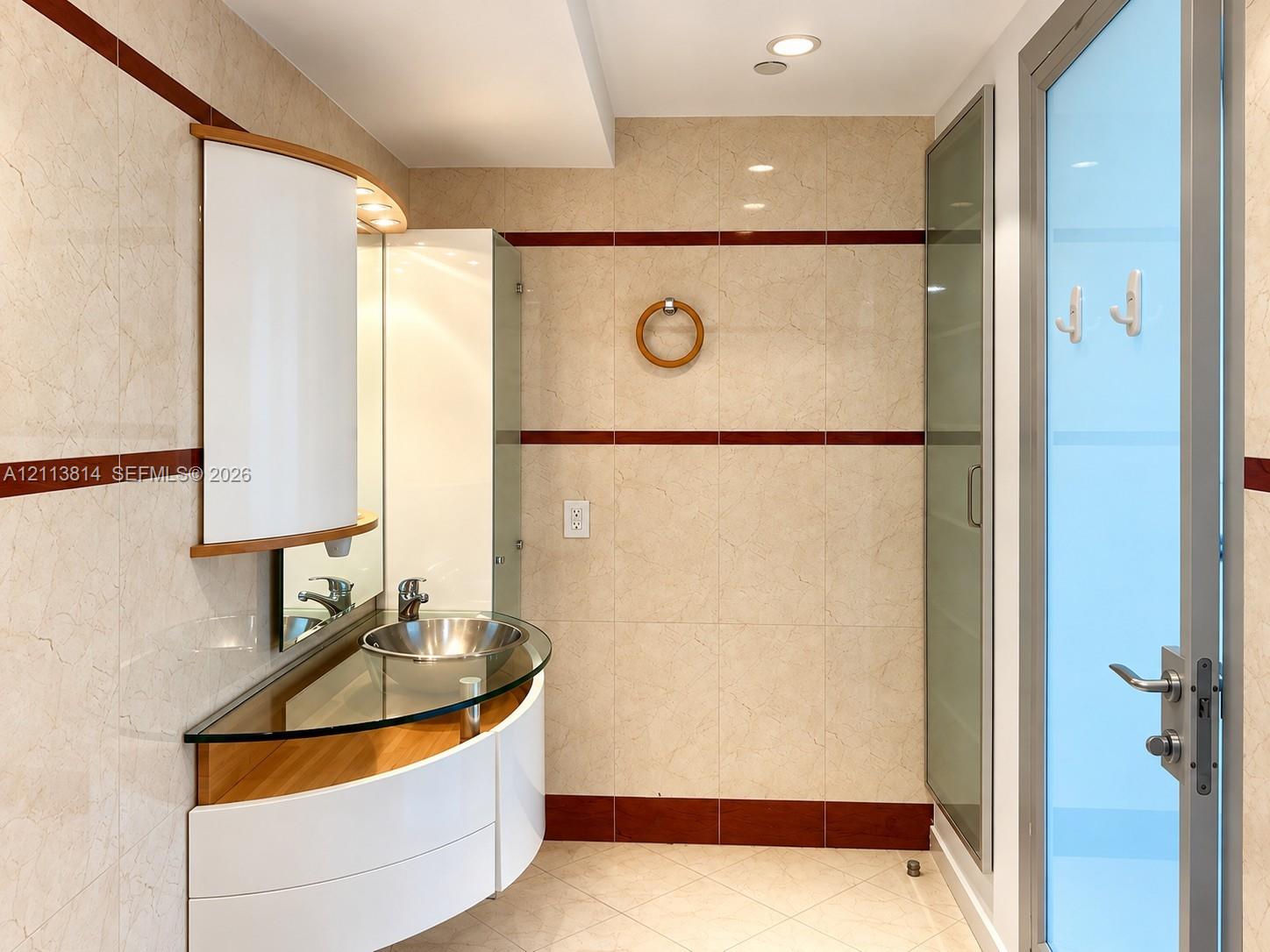 Curved floating vanity featuring a glass countertop, metallic vessel sink, and white cabinet storage, finished with full-height tile walls and recessed lighting