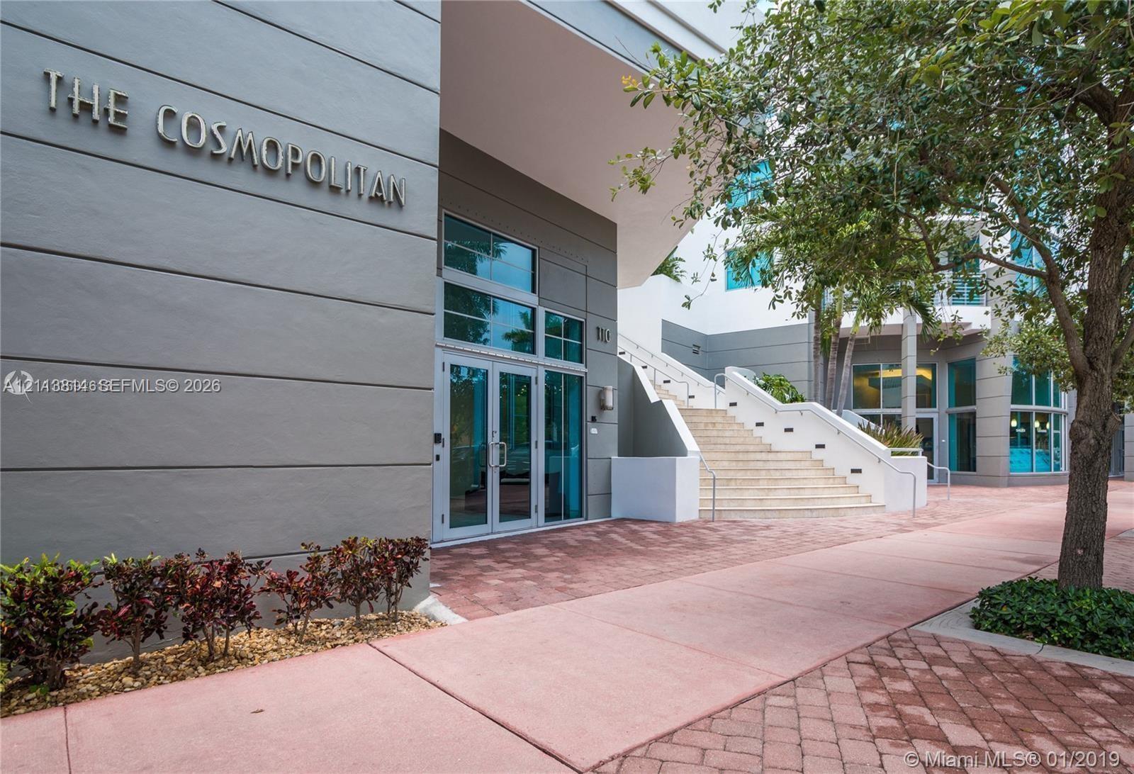 Building entrance featuring a multi-story facade, glass-paned double doors, stone staircase with metal railings, and patterned paver walkway