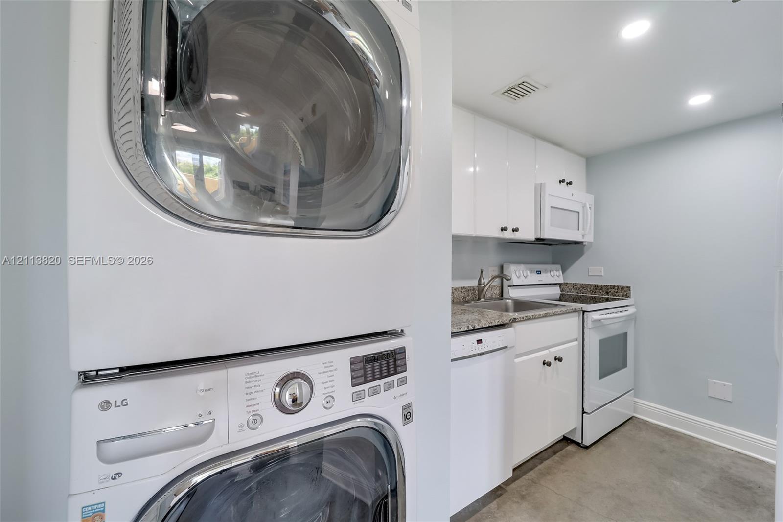 Laundry area featuring a stackable washer and dryer, white cabinetry, stone-finish countertops, stainless steel sink with a high-arch faucet, and white appliances Laundry area featuring a stackable washer and dryer, white cabinetry, stone-finish countertops, stainless steel sink with a high-arch faucet, and white appliances