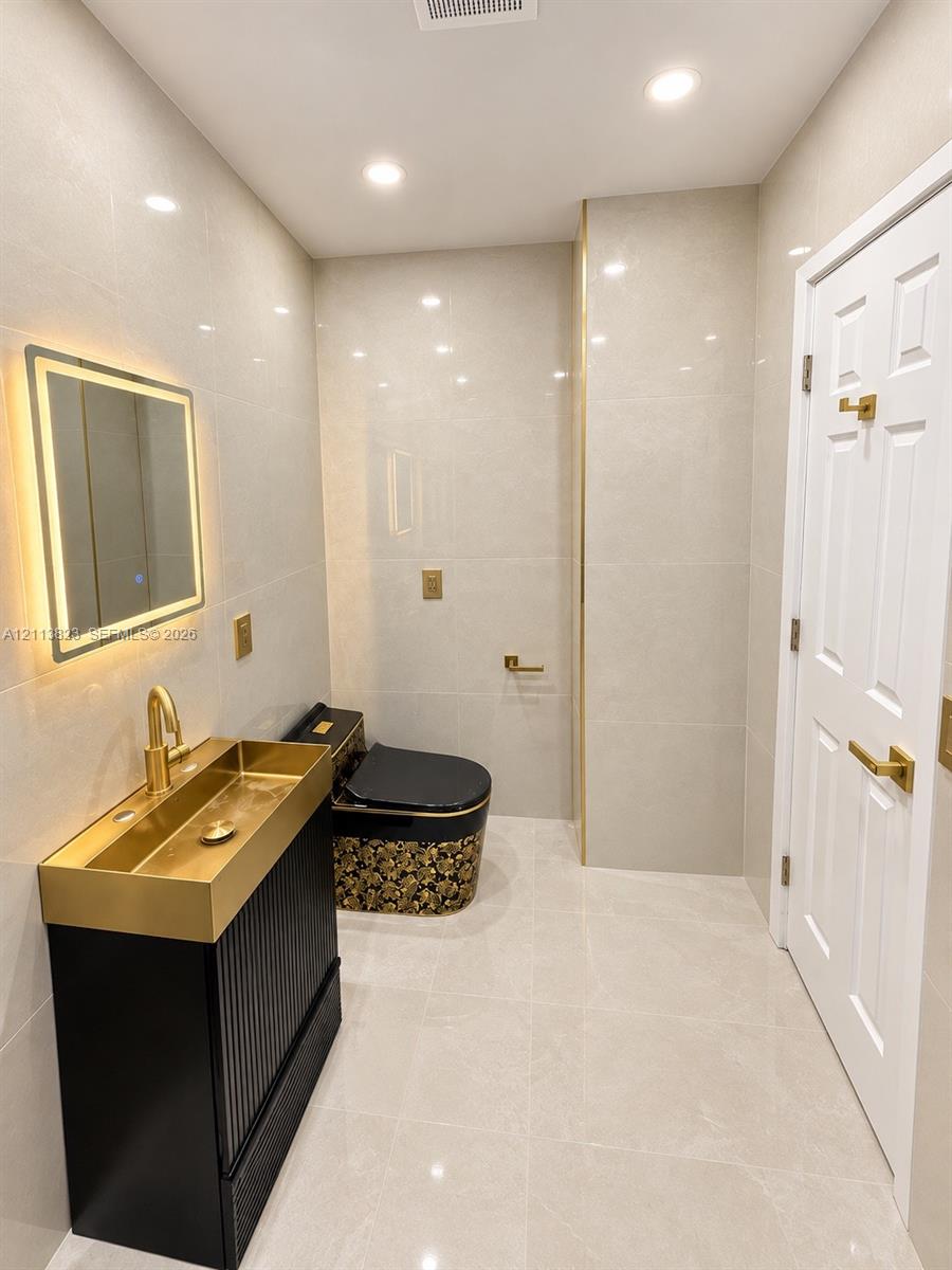 Modern vanity featuring a metallic basin, wall-mounted illuminated mirror, patterned decorative toilet, recessed ceiling lighting, and floor-to-ceiling tilework