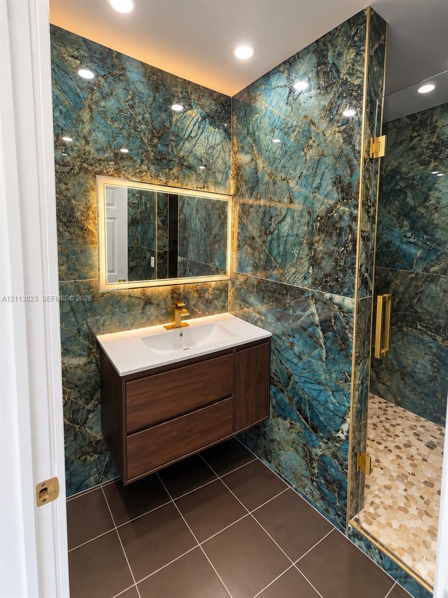 Floating wood-finish vanity with an integrated sink, polished gold-tone faucet, backlit wall mirror, stone-finish wall cladding, and walk-in shower with gold-tone hardware