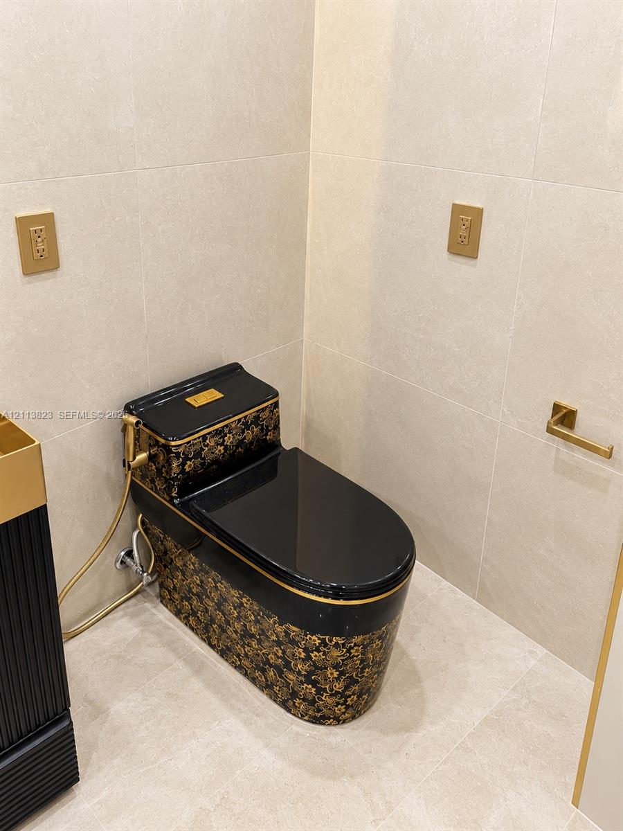 One-piece patterned toilet featuring a black and gold floral finish, complemented by gold-tone hardware, a wall-mounted toilet paper holder, and large-format neutral wall tiles