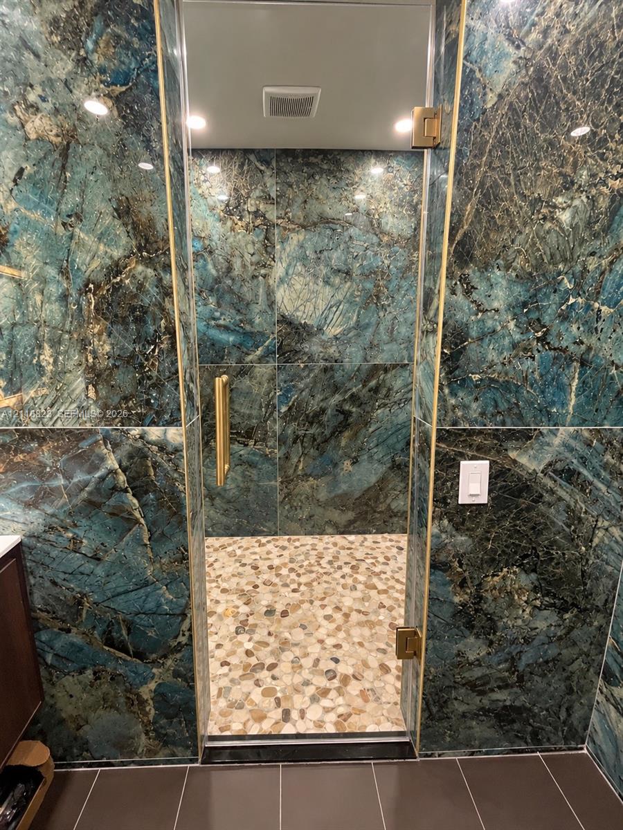 Shower enclosure featuring large-format polished stone wall tile in blue and green tones, pebble-style floor tile, and gold-finish hardware