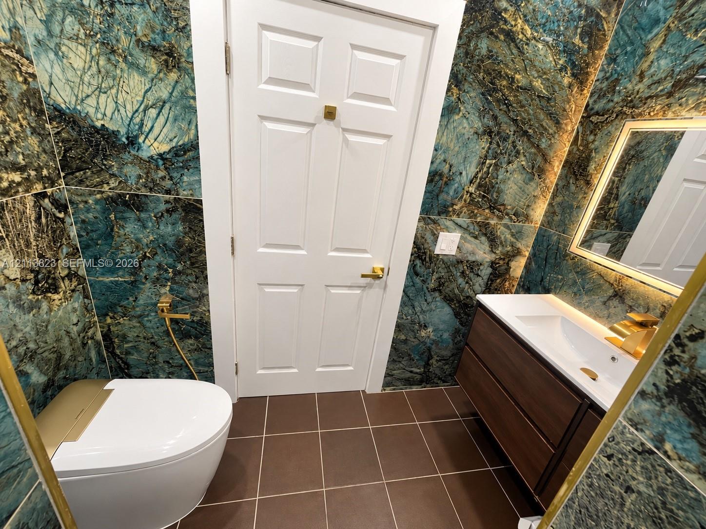 Full-height dark stone-patterned wall tiling, floating vanity with integrated rectangular sink, gold-tone hardware, backlit wall mirror, and modern low-profile toilet