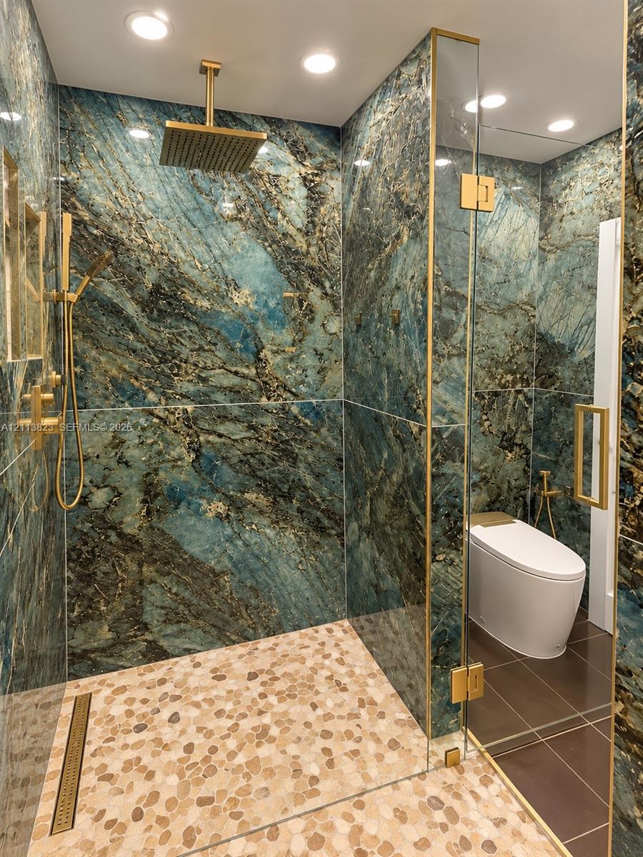 Walk-in shower featuring floor-to-ceiling stone slab wall surfacing, pebble-stone shower flooring, gold-finish rain shower head with handheld attachment, and frameless glass enclosure with gold-tone hardware