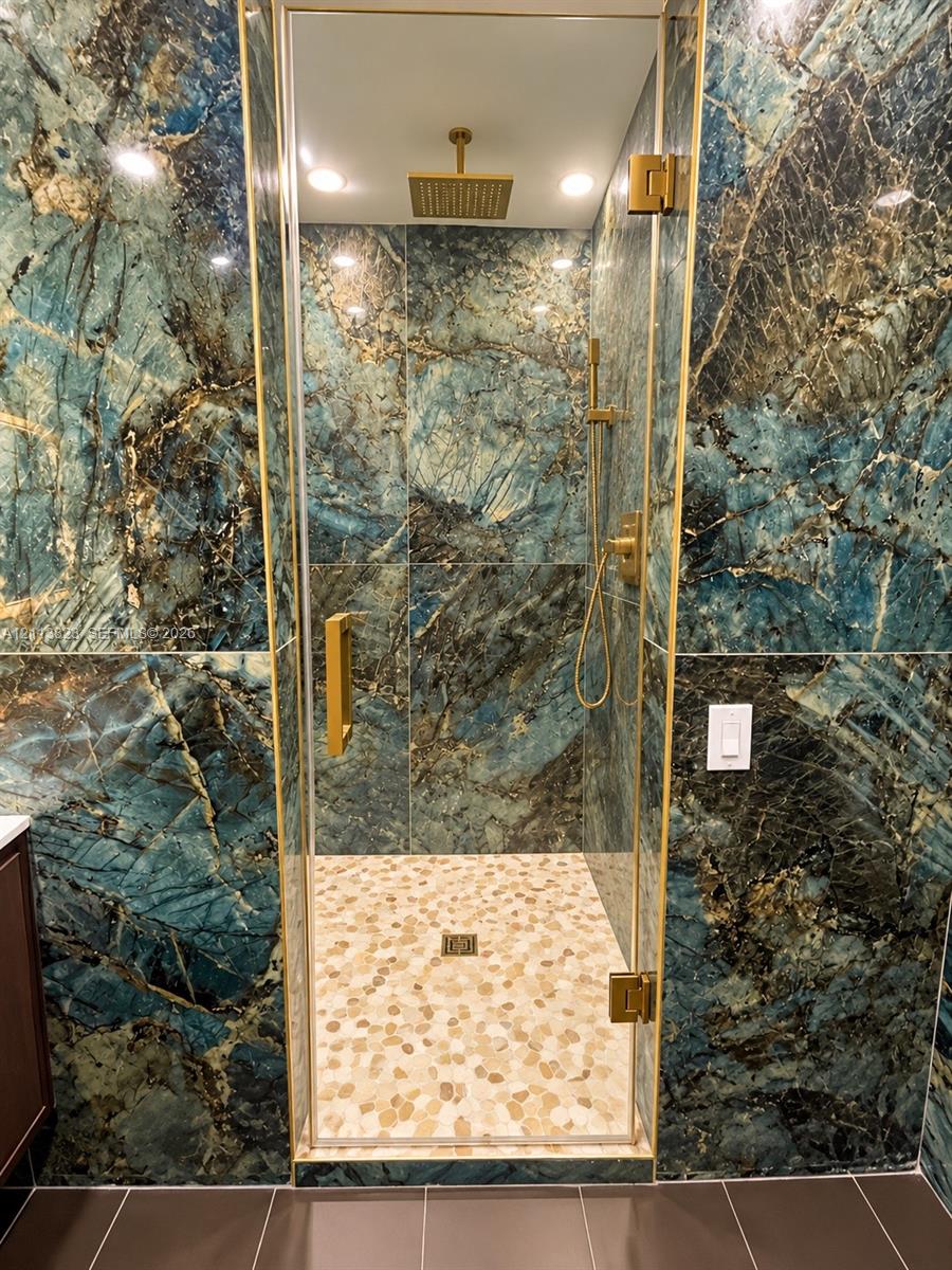Walk-in shower featuring large-format stone-finish wall tile, frameless glass enclosure, gold-tone hardware, rainfall showerhead, and a pebble-stone floor finish
