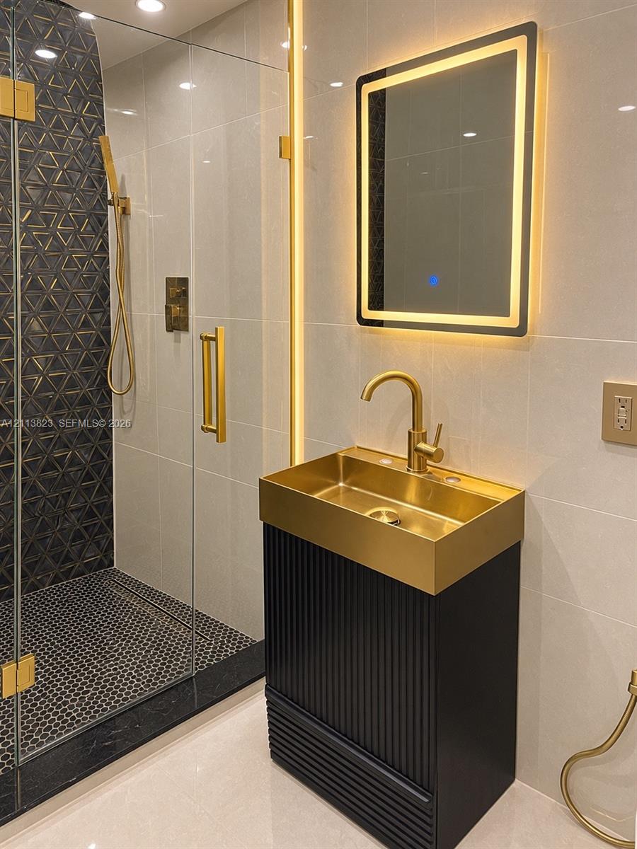Custom vanity featuring a fluted dark cabinet base, integrated metallic sink, and gold-tone hardware, paired with a glass-enclosed shower, geometric-patterned tile, and LED-backlit mirror